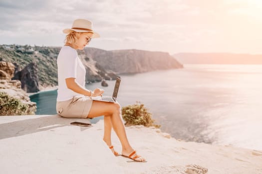 Successful business woman in yellow hat working on laptop by the sea. Pretty lady typing on computer at summer day outdoors. Freelance, travel and holidays concept.