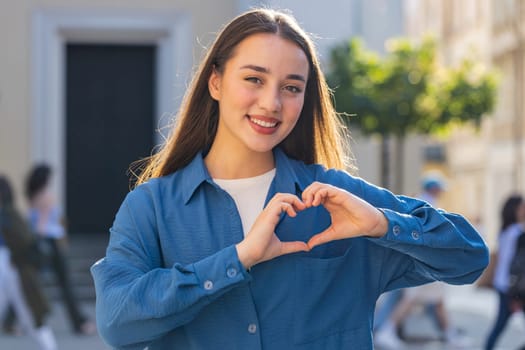 I love you. Yowoman makes symbol of love, showing heart sign to camera, express romantic feelings, express sincere positive feelings. Charity, gratitude, donation. Outdoors in sunny city street