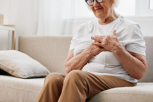 Elderly woman severe chest pain sitting on the sofa, health problems in old age, poor quality of life. Grandmother with gray hair holds her chest with her hands, women's health, breast cancer. High quality photo