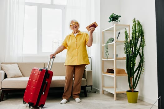 Happy senior woman with passport and travel ticket packed a red suitcase, vacation and health care. Smiling old woman joyfully stands in the house before the trip. High quality photo