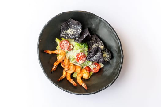 Exquisite serving Caesar salad with shrimps on a black plate on a white background