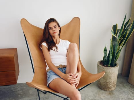 Woman sitting in leather armchair smile with teeth Lifestyle in white T-shirt and blue jean shorts, relaxing at home stylish modern interior with white walls, close-up. High quality photo