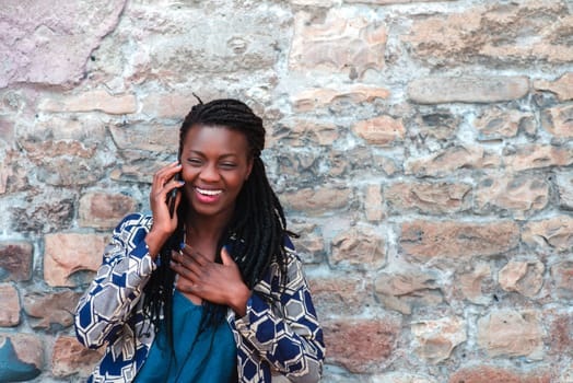 confident African young adult woman holding cell phone calling , receives good news, smiling, happy, outdoors in the village over a wall.