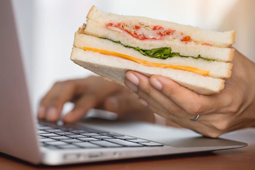 Hand holding a delicious sandwich and using laptop computer for eating while working concept.