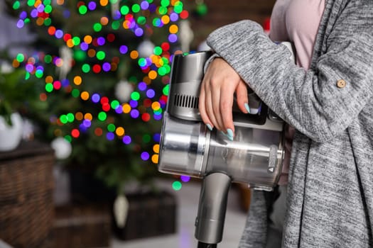 A short break during household cleaning. A girl leaning against a vacuum cleaner against a Christmas tree dressed with Christmas lights and other decorations.