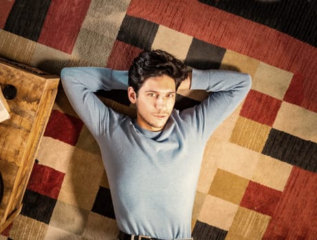Cool dark-haired handsome young man laying on the floor over colorful rug, looking at camera, hand under his head