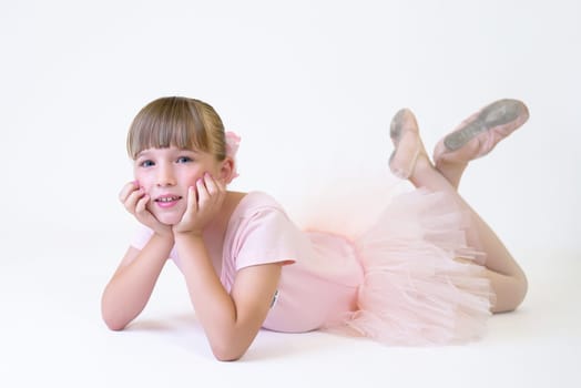 Little ballerina dancer in a pink tutu academy student posing on white background. Dancing girl lying on the floor posing for the camera.