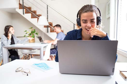 enterprising girl smiling with headphones working with the computer on a white desk in a space shared with other workers in the background