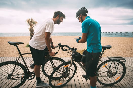 Cyclists on a bike on a wooden walkway on the beach with the sea and a bridge in the background looking at the mobile phone. Clear sky background with copyspace. sport concept. urban cycling concept. technology concept