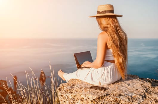 Successful business woman in yellow hat working on laptop by the sea. Pretty lady typing on computer at summer day outdoors. Freelance, travel and holidays concept.