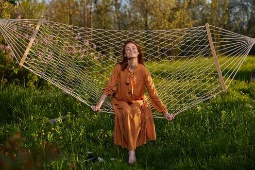 horizontal photo of a beautiful, red-haired woman lying in a hammock enjoying a rest in a long orange dress, on a warm summer day, smiling happily looking at the camera. High quality photo