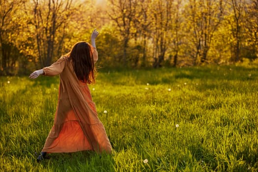 a slender woman with long hair stands in a field with her back to the camera, illuminated by the rays of the setting sun and happily poses enjoying the warm weather and rest. High quality photo