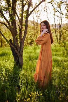 a beautiful woman stands in a long orange dress, in the countryside, near a flowering tree, during sunset, and holds her hand near her face. Vertical photography. High quality photo
