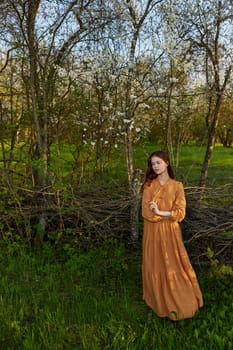 an elegant, sophisticated woman poses relaxed standing near a wicker fence at the dacha in a long orange dress looking to the side. Vertical photography in nature. High quality photo