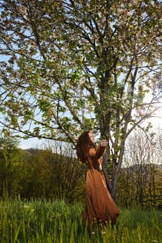 a slender, attractive woman with long red hair is standing in the countryside near a flowering tree in a long orange dress and happily spinning holding the dress. Vertical photography. High quality photo