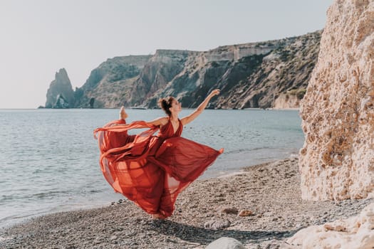 Woman red dress sea. Female dancer in a long red dress posing on a beach with rocks on sunny day. Girl on the nature on blue sky background