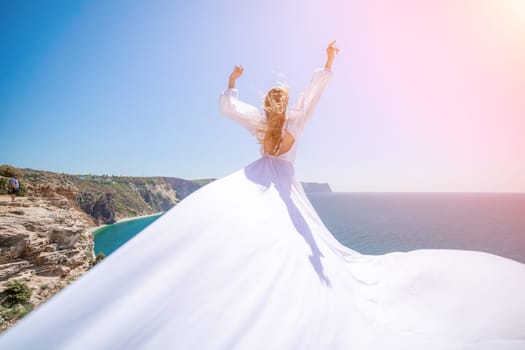 woman sea white dress. Blonde with long hair on a sunny seashore in a white flowing dress, rear view, silk fabric waving in the wind. Against the backdrop of the blue sky and mountains on the seashore