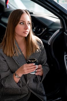 Happy woman coffee. she stands next to the car in the underground parking. Dressed in a gray coat, holding a glass of coffee in her hands, a black car