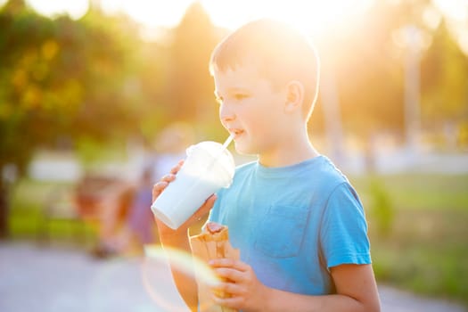 Child boy eats a hot-dog sandwich and drinks milkshake the park at sunset outdoors.