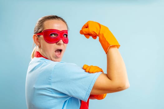 Confidence superhero house cleaner showing her powerful muscle arm with wearing red apron and eye mask on blue background