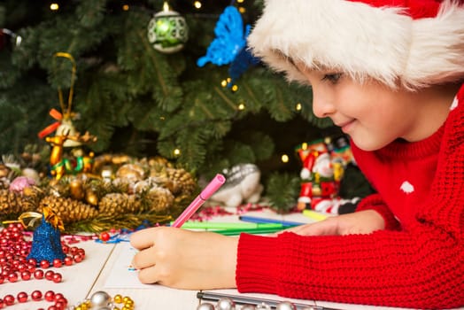 Little boy in Santa hat writes letter to Santa Claus. Kid in Santa hat writing wish list
