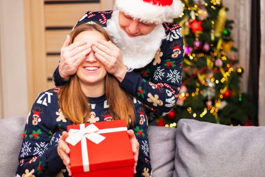 Man dressed as Santa covering his girlfriend's eyes while gives her Christmas surprise. Man giving christmas present to his girlfriend.