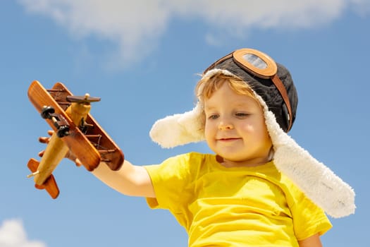 Happy child boy playing with wooden toy airplane on blue sky background with clouds.