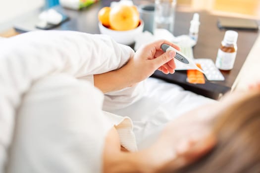 Sick young woman covered with a blanket lying in bed with high fever and a flu. A lot of medicines on table in bedroom.