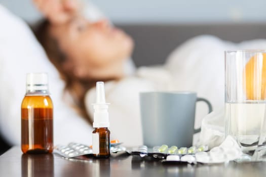 Sick young woman covered with a blanket lying in bed with high fever and a flu. A lot of medicines on table in bedroom.