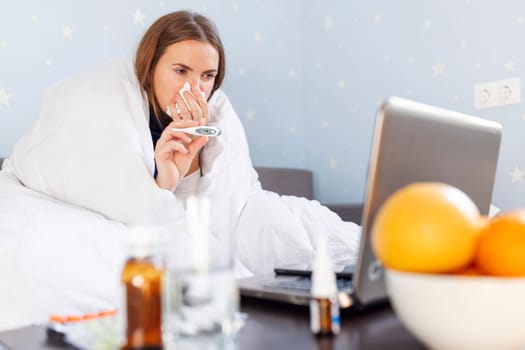 Sick young woman with bad cold, flu or coronavirus fever holding thermometer and talking to telemedicine service doctor. Sick woman having online medical consultation video call.