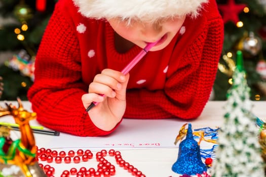 Little child is writing letter to Santa Claus at table. A kid in Santa hat is writing a letter to Santa.