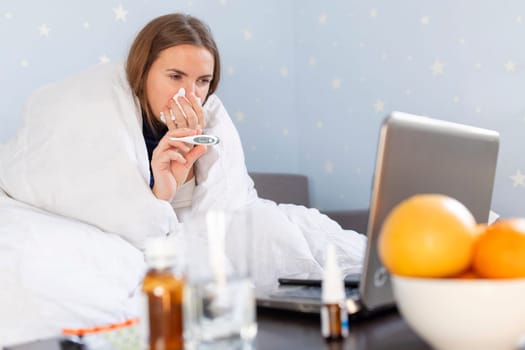 Sick young woman with bad cold, flu or coronavirus fever holding thermometer and talking to telemedicine service doctor. Sick woman having online medical consultation video call.