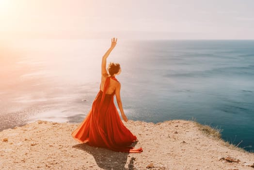 Side view a Young beautiful sensual woman in a red long dress posing on a rock high above the sea during sunrise. Girl on the nature on blue sky background. Fashion photo.