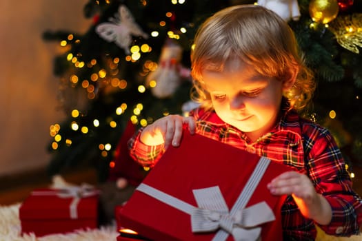 Cute little boy opening magic Christmas gift by a Christmas tree in cozy living room in winter. Merry Christmas