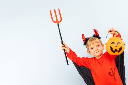 Happy Halloween. Cute little boy in devil halloween costume with trident and pumpkin basket jack-o-lantern on light blue background.