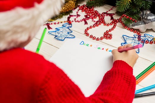 Little child is writing letter to Santa Claus at table. A kid in Santa hat is writing a letter to Santa.
