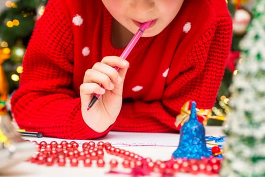 Little child writes letter to Santa. Cute boy in Santa hat writes a letter to Santa near the Christmas tree.