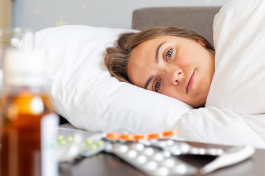 Sick young woman covered with a blanket lying in bed with high fever and a flu. A lot of medicines on table in bedroom.