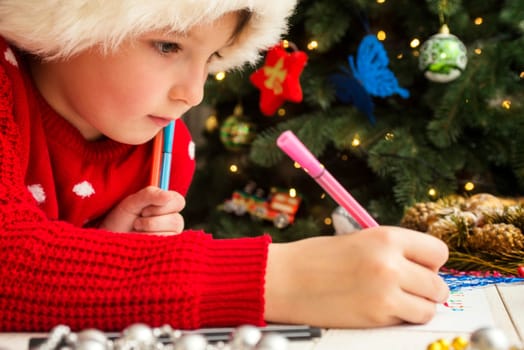 Little child is writing letter to Santa Claus at table. A kid in Santa hat is writing wishlist near the Christmas tree.