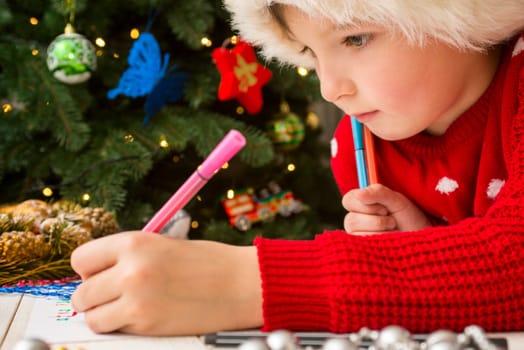 Little boy in Santa hat writes letter to Santa Claus. Kid in Santa hat writing wish list