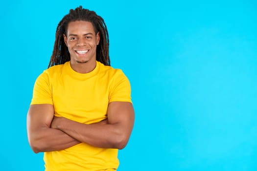 Happy latin man with dreadlocks standing with arms crossed in studio with blue background