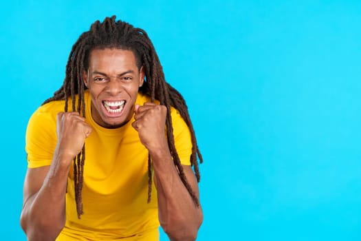 Happy hispanic man with dreadlocks gesturing with fists while celebrating in studio with blue background