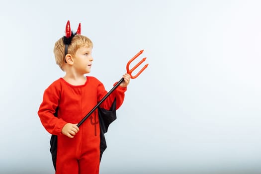 Happy Halloween. Cute little boy in devil halloween costume with horns and trident on light blue background.