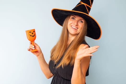 Happy Halloween. Happy brunette woman in halloween witch costume with black hat holding glass of cocktail on a light background. Halloween party.