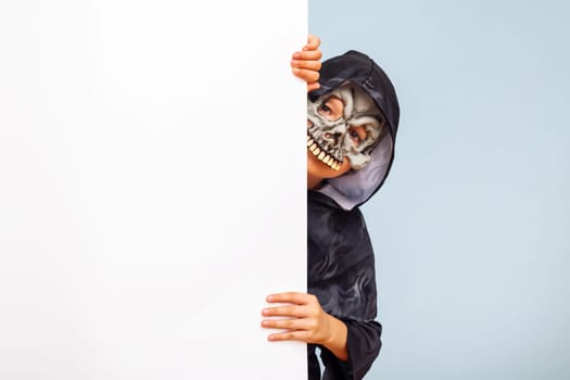 Happy Halloween. Cute little boy in a costume hiding behind white banner blank. Happy kid holding cardboard background with copy space.