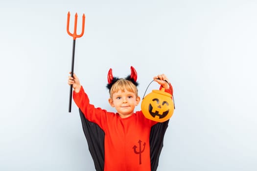 Happy Halloween. Cute little boy in devil halloween costume with trident and pumpkin basket jack-o-lantern on light blue background.