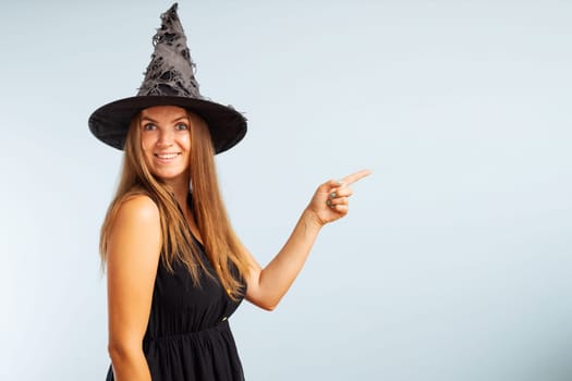 Woman in witch costume pointing with finger at copyspace side and looking at camera on blue background. Happy Halloween.