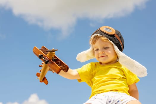 Happy child boy playing with wooden toy airplane on blue sky background with clouds.