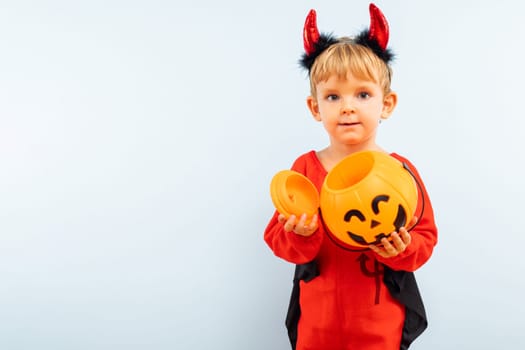 Happy Halloween. Cute little boy in devil halloween costume with pumpkin basket jack-o-lantern on light blue background.