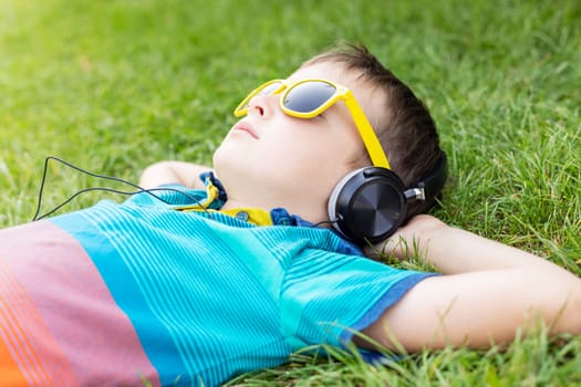 Happy boy in sunglasses lying on the grass in the park and listening to music in headphones on a sunny day.